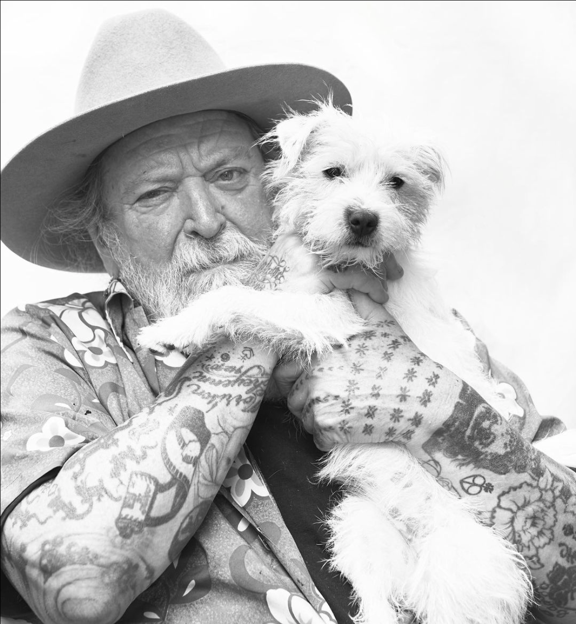 Henk Schiffmacher, a famous tattoo master, holding his terrier dog, Baby. Both are looking directly at the camera with a serene expression.